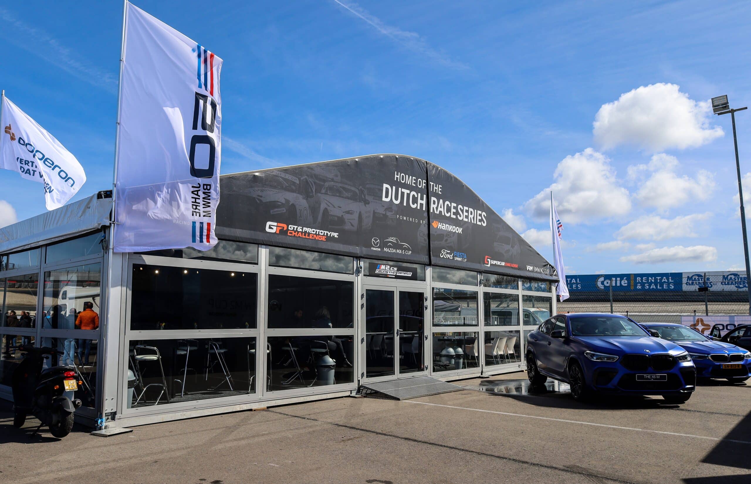 Aluhal waar groot 'Home of the Dutch race series' op staat. Links van de Aluhal staat een grote witte vlag van BMW en aan de rechterkant staan twee blauwe BMW's. Om de Aluhal heen bevind zich een racebaan